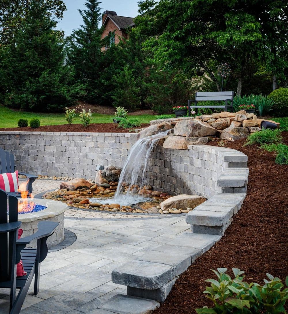 Outdoor landscape with a stone waterfall, seating area, and lush greenery.