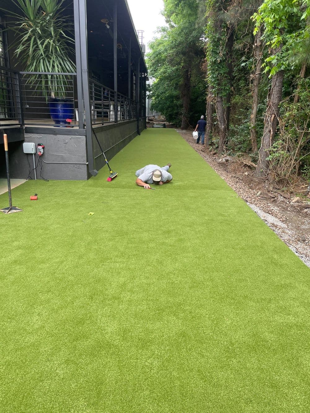 Person installing artificial turf along a lush green pathway next to a building.