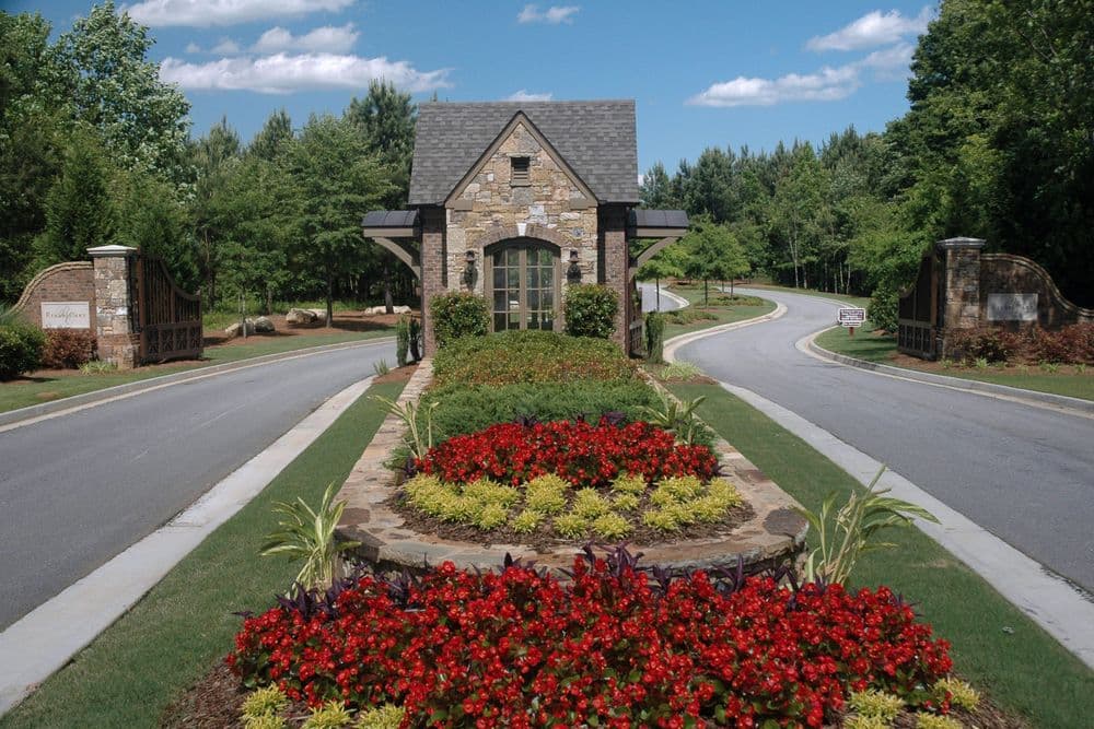 Stone entrance gate with flower beds and trees, leading to a scenic neighborhood road.